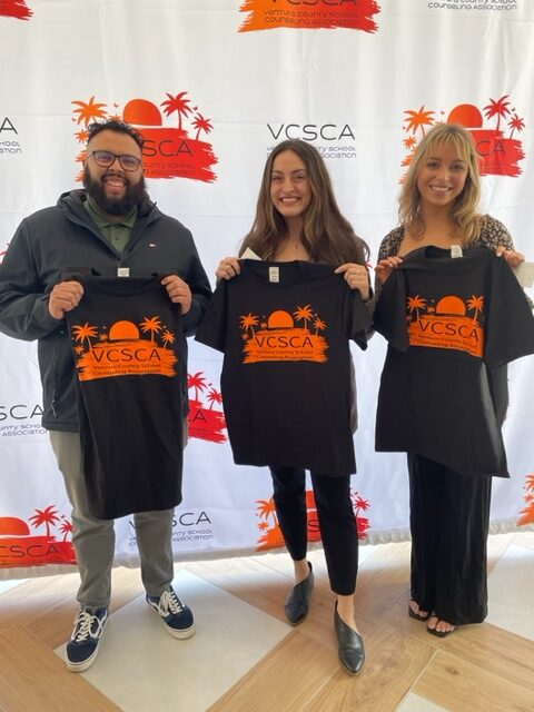 This photo shows three people standing in front of a white backdrop, holding black t-shirts with the VCSCA logo in orange. The individuals are VCSCA scholarship recipients, and from left to right, the group includes a bearded brunette man, a brunette woman, and a blonde woman. The VCSCA logo is visible all over the white backdrop behind the group.
